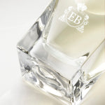 Detail of the bottom of the 100 ml bottle, with transparent glass and yellowish perfum. Detail of logo with the EB initials in white ink. Fabulous Magnolia, a fragrance by Eric Butherbaugh.