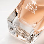 Detail of the bottom of the 100 ml bottle, with transparent glass and orangey perfum. Detail of logo with the EB initials in white ink. Maiden Orange Blossom, a fragrance by Eric Butherbaugh.