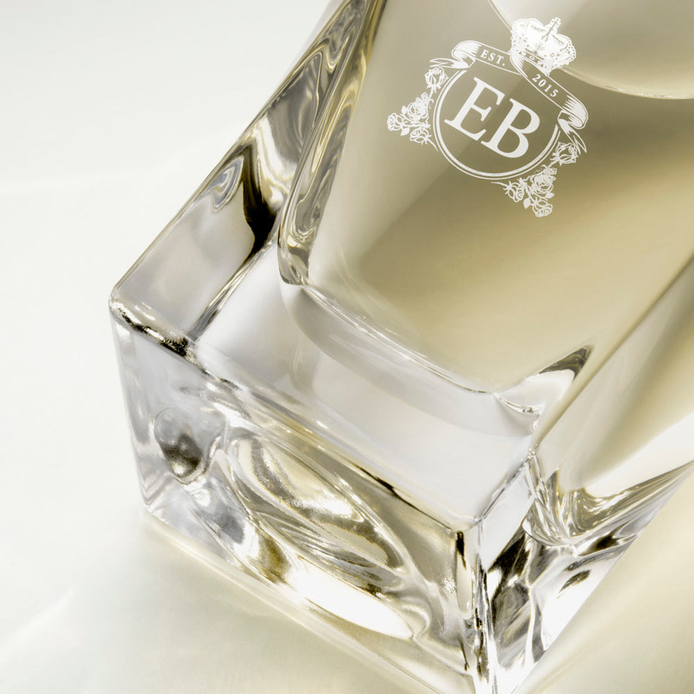 Detail of the bottom of the 100 ml bottle, with transparent glass and yellowish perfum. Detail of logo with the EB initials in white ink. Virgin Lily of the Valley, a fragrance by Eric Butherbaugh.