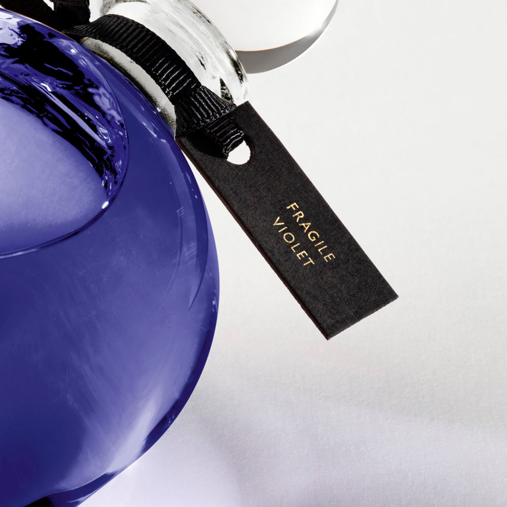 Detail of the top of the 250 ml bottle, with transparent glass and violet perfum. Spherical cap with black ribbon. Fragile Violet, a fragrance by Eric Butherbaugh.