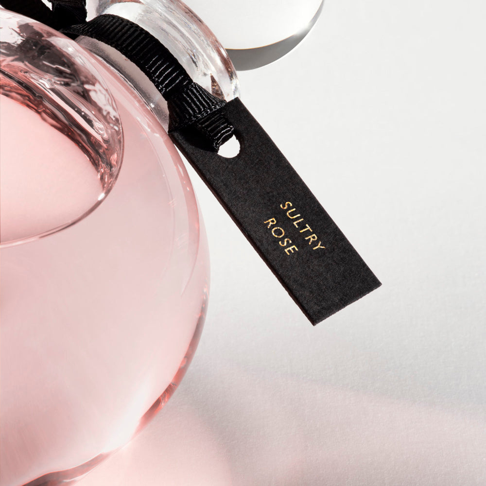 Detail of the top of the 250 ml bottle, with transparent glass and pinkish perfum. Spherical cap with black ribbon. Sultry Rose, a fragrance by Eric Butherbaugh.