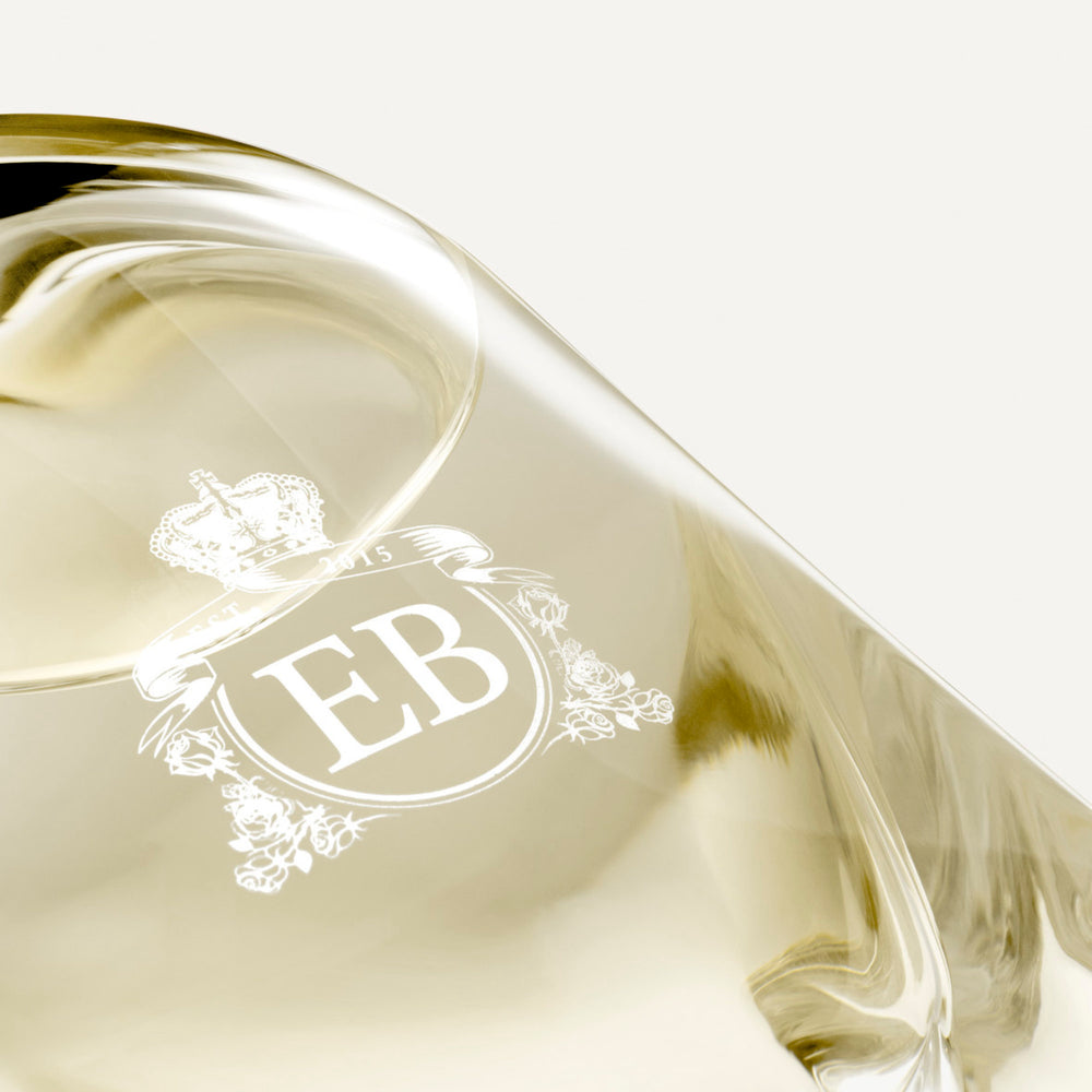 Detail of the bottom of the 250 ml bottle, with transparent glass and yellowish perfum. Detail of logo with the EB initials in white ink. Virgin Lily of the Valley, a fragrance by Eric Butherbaugh.